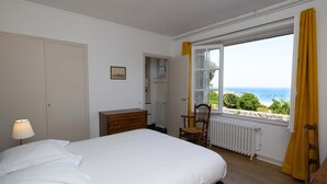 A bedroom with a bed, a dresser, a chair, a window with a view of the sea, and a radiator. - Renovated villa in park, very large sea view, 5 minutes walk from the beach (Saint-Cast-le-Guildo)