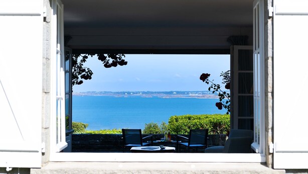 A view from a window overlooking a body of water, with a table and chairs outside. - Renovated villa in park, very large sea view, 5 minutes walk from the beach (Saint-Cast-le-Guildo)