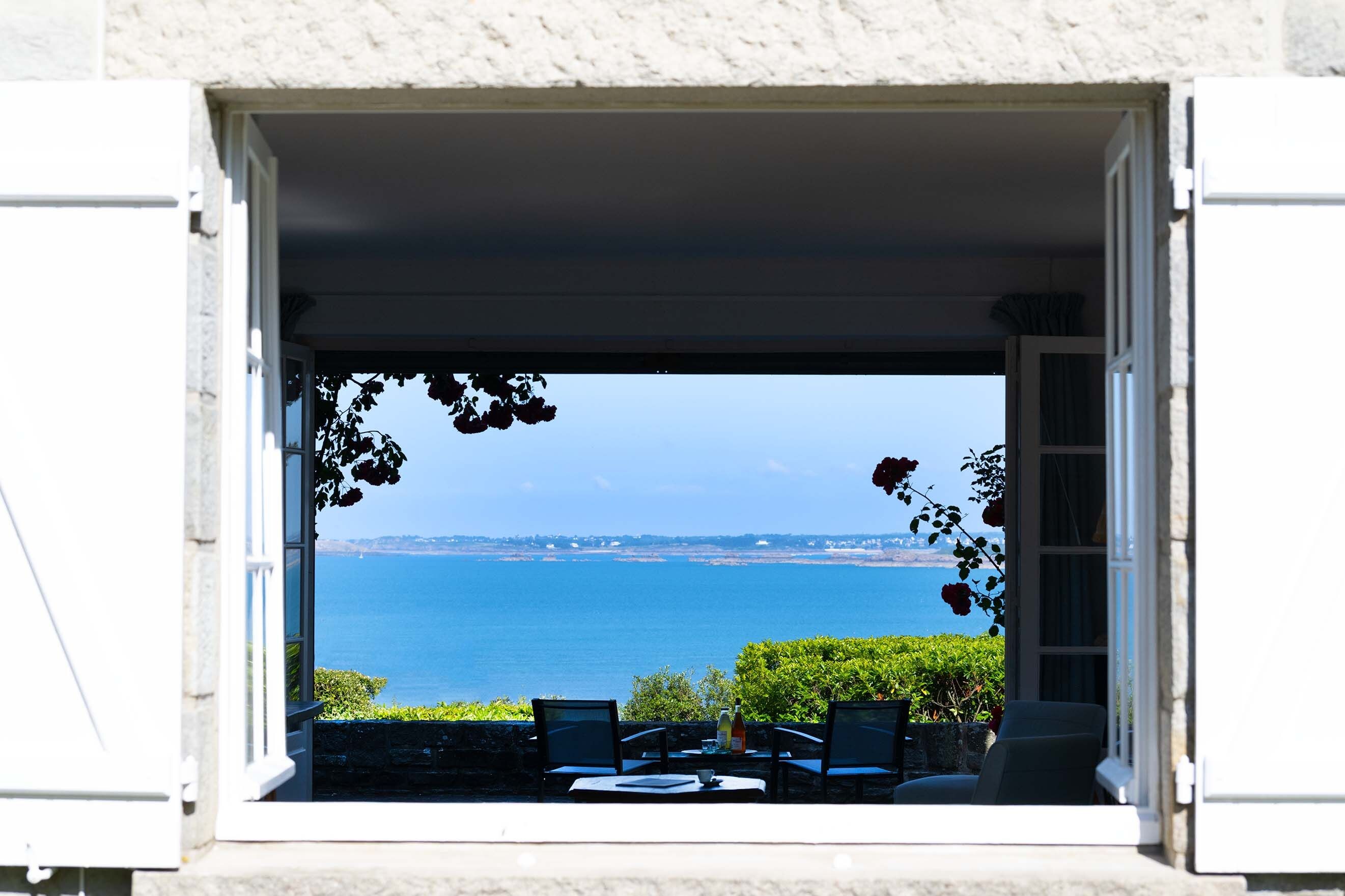 Une vue depuis une fenêtre donnant sur une étendue d’eau, avec une table et des chaises à l’extérieur.