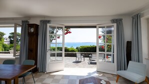 A room with a checkered floor, a wooden table, a white armchair, and a view of the sea through large glass doors. - Renovated villa in park, very large sea view, 5 minutes walk from the beach (Saint-Cast-le-Guildo)
