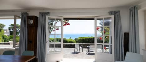 A room with a checkered floor, a wooden table, a white armchair, and a view of the sea through large glass doors.
