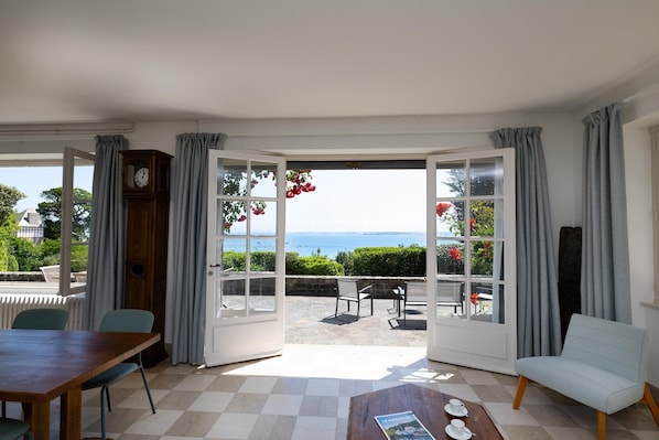 A room with a checkered floor, a wooden table, a white armchair, and a view of the sea through large glass doors. - Renovated villa in a park, spectacular ocean view, 5 minutes to the beach (Saint-Cast-le-Guildo)