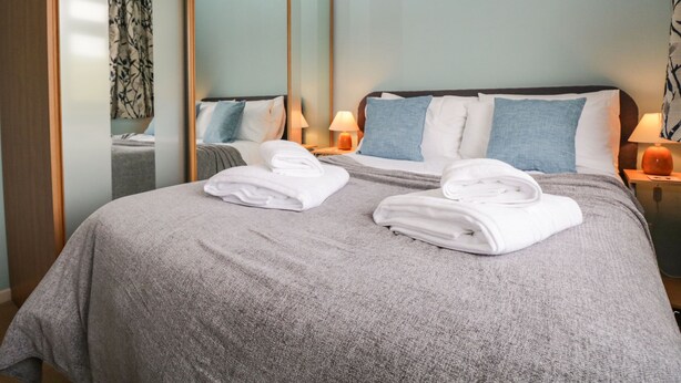 A hotel room with a large bed, two bedside tables with lamps, a mirror, and a view of a room with a patterned curtain. - Marsh Garth (Kirkby-in-Furness)