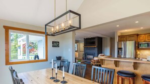 A modern kitchen with a central island, a dining area with a wooden table and chairs, and a large window with a view of the outdoors. - The Summit | Nature Lovers Dream! Family Friendly (Vernon)