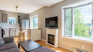 A modern living room with a sofa, a coffee table, and a fireplace. There is a kitchen area with cabinets and a refrigerator. A television is mounted above the fireplace.