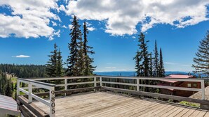 A wooden deck with a white railing, overlooking a forested area and distant mountains under a partly cloudy sky.