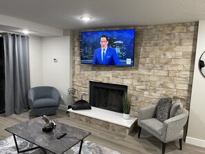 Een moderne woonkamer met een stenen open haard, een flatscreen televisie, een grijze fauteuil, een gestreept fauteuil en een marmeren salontafel.