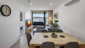 A modern living room with a dining area, a large window with a view, a clock, and an air conditioning unit. - Bay View 2-Bedroom Apartment (Batemans Bay)