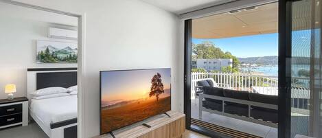 A modern hotel room with a bed, a TV, a balcony with a view of the sea, and a dining area.