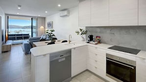 A modern kitchen with a dishwasher, oven, and a dining area with a view. - Bay View 2-Bedroom Apartment (Batemans Bay)