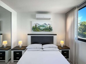 Une chambre d’hôtel comprenant un lit, deux tables de chevet avec des lampes, un climatiseur fixé au mur, un tableau représentant un paysage de montagne et une grande fenêtre avec des rideaux.