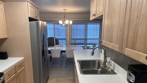 A modern kitchen with a stainless steel refrigerator, a double sink, and a coffee maker. There is a dining area with a table and chairs, and a view of the ocean through large windows. - The Flathead Overlook at Lakeplace (Polson)