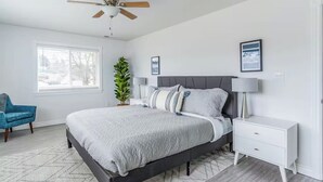 A modern bedroom with a bed, bedside tables, a ceiling fan, a window, and a plant. - The Flathead Overlook at Lakeplace (Polson)