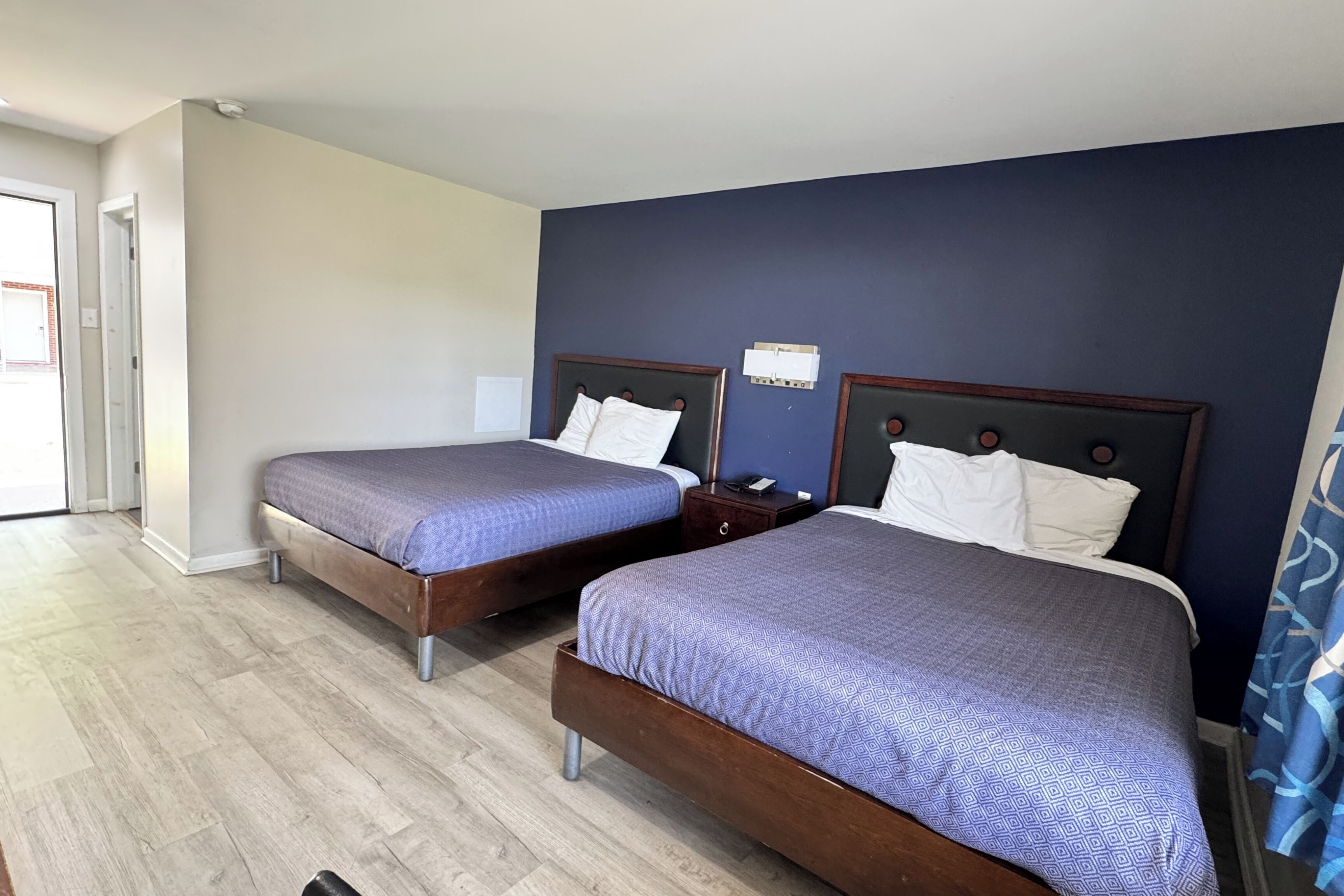 A hotel room with two beds, a wooden floor, a dark blue accent wall, and a visible electrical outlet.