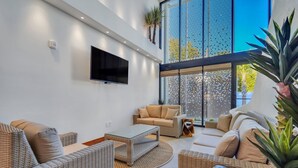 A modern living room with wicker furniture, a flat-screen TV, and large windows offering a view of the outdoors. - Vacation Villa 5 mins to King ‘The Royal Palm’ (Charleston)