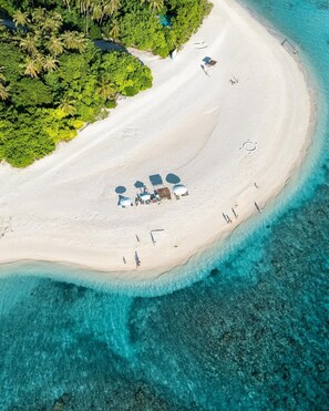 Vue aérienne d’une plage aux eaux turquoise et limpides, bordée d’un rivage sablonneux et ponctuée de quelques parasols et de chaises longues.