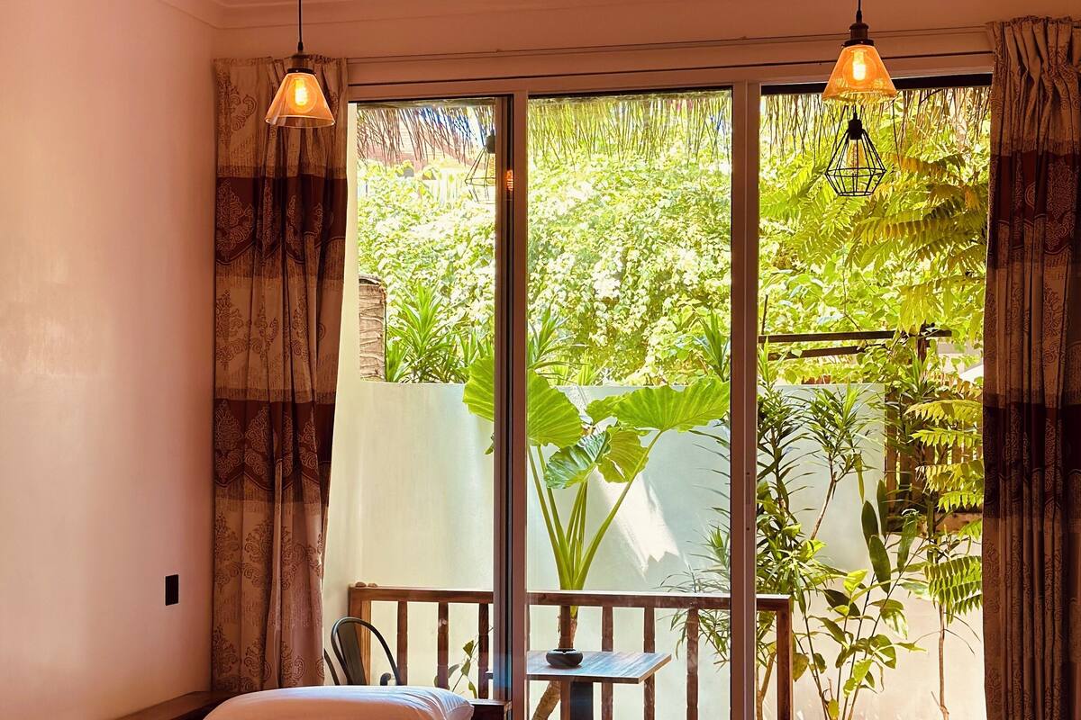 A room with a bed, a small table, and a chair. There are curtains on the windows, and a plant is visible outside.