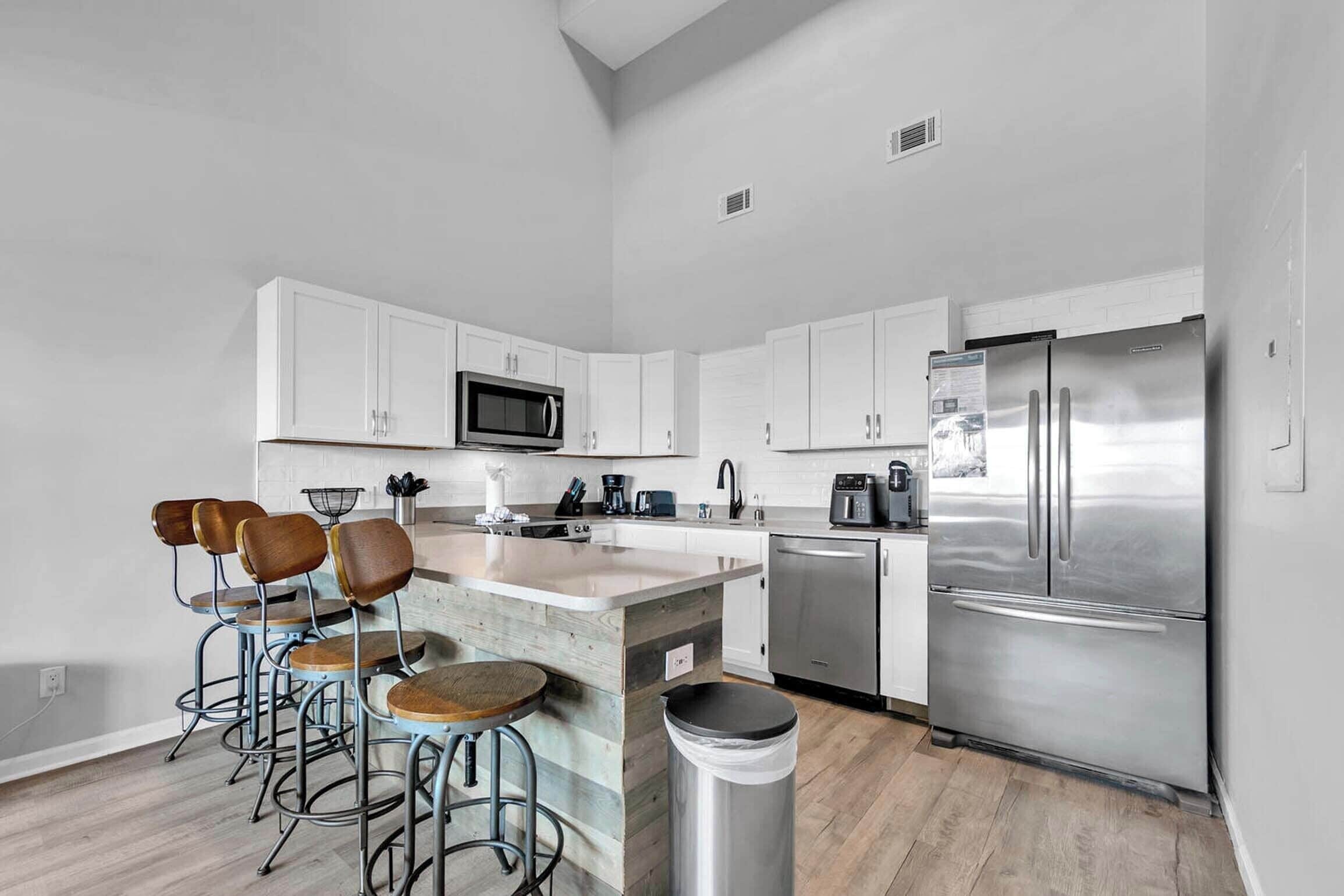 A modern kitchen with white cabinets, a stainless steel refrigerator, a microwave, and a coffee maker. There are three bar stools with wooden seats and metal frames. A trash can is visible in the foreground.