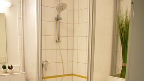 A corner shower with a glass door, a white toilet, and a sink with a blue trash bin. - Posthus Apartment 201, close to the beach and central (Borkum)