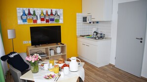 A room with a yellow wall, a painting, a TV, a kitchenette, and a breakfast setup on a table. - Posthus Apartment 201, close to the beach and central (Borkum)