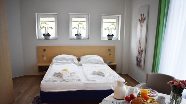 A hotel room with a double bed, two bedside tables, a small table with breakfast items, and three windows with potted plants. - Posthus Apartment 201, close to the beach and central (Borkum)