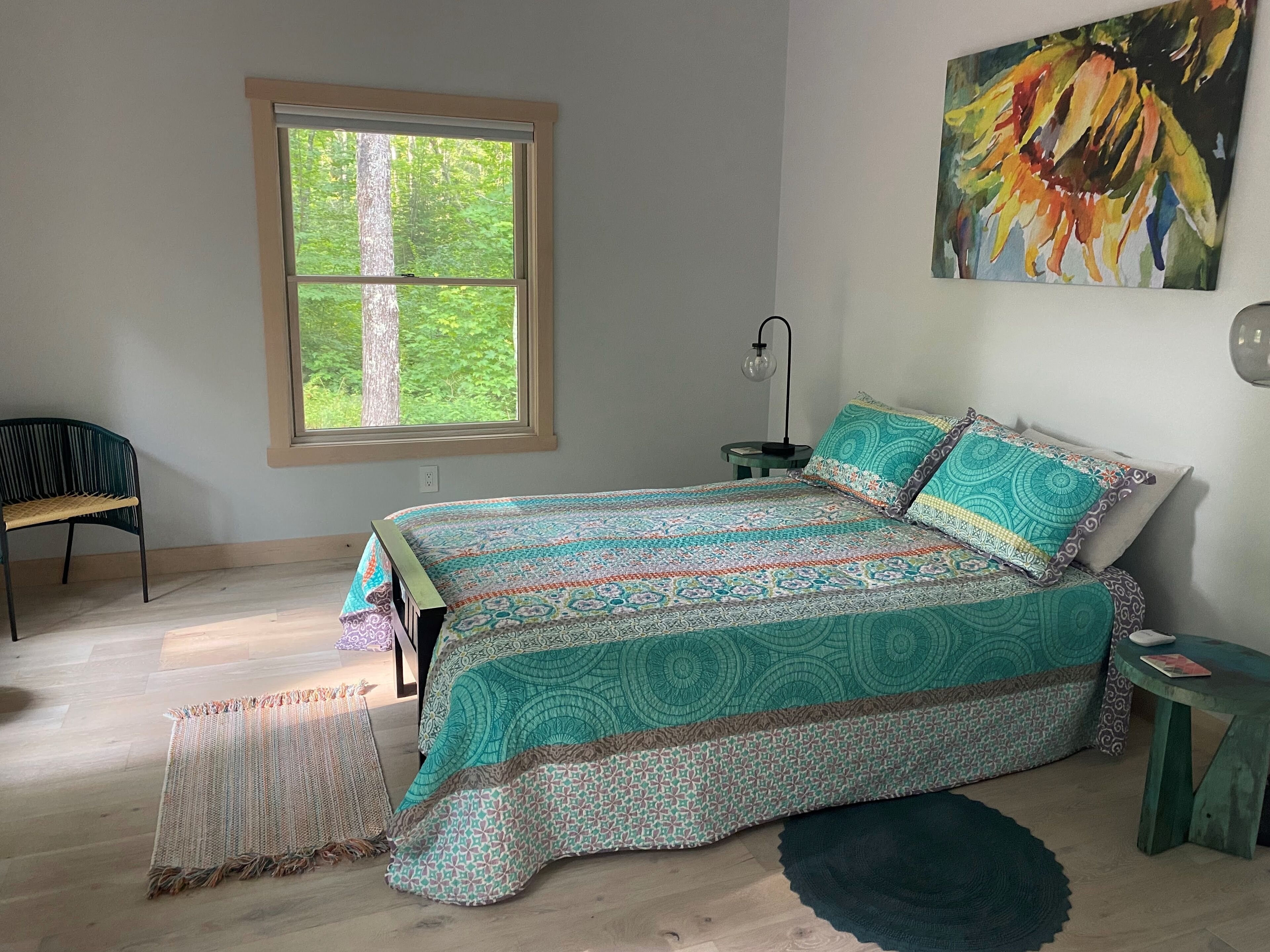 Un dormitorio con una cama grande, una ventana que da a unas copas de árboles, una silla, una mesita y un cuadro en la pared.