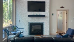 A living room with a stone fireplace, a flat-screen TV mounted above it, and a large window offering a view of trees.
