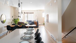 Un espace de salle à manger moderne, avec une longue table dressée pour six personnes, entourée de chaises noires. Des suspensions lumineuses sont placées au-dessus de la table, et un canapé avec des coussins se trouve à l’arrière-plan.