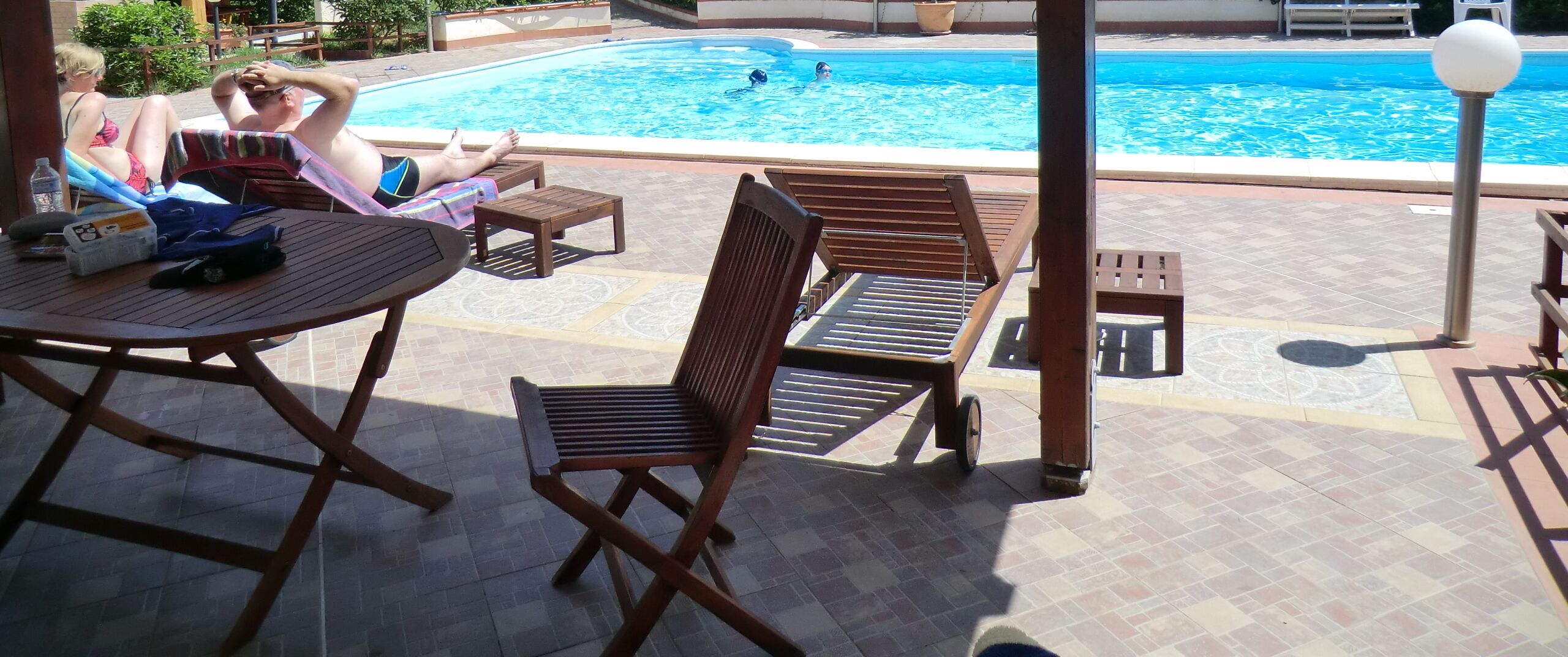 Un espace aménagé au bord de la piscine, avec des chaises longues et une table, une piscine où des personnes nagent, et une vue dégagée sur la piscine.