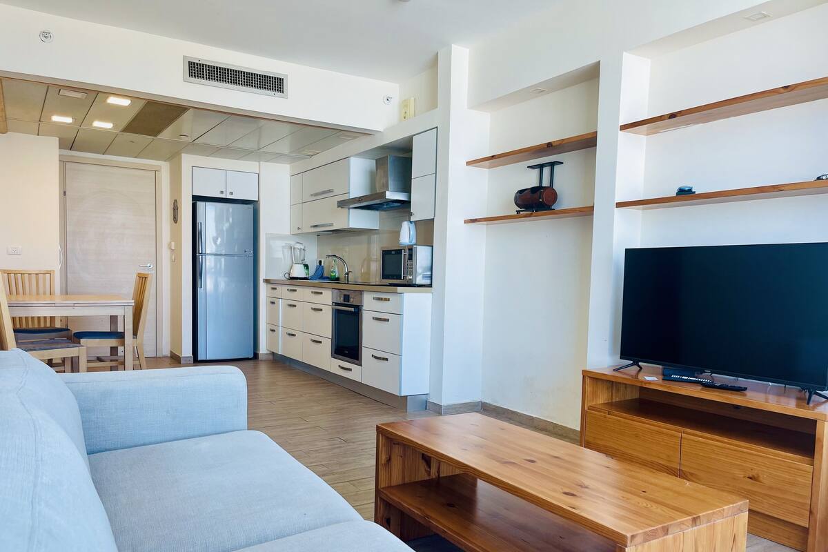 A modern kitchen with white cabinets, a built-in oven, and a microwave. There is a dining area with a wooden table and chairs, and a living room with a sofa and a wooden coffee table.