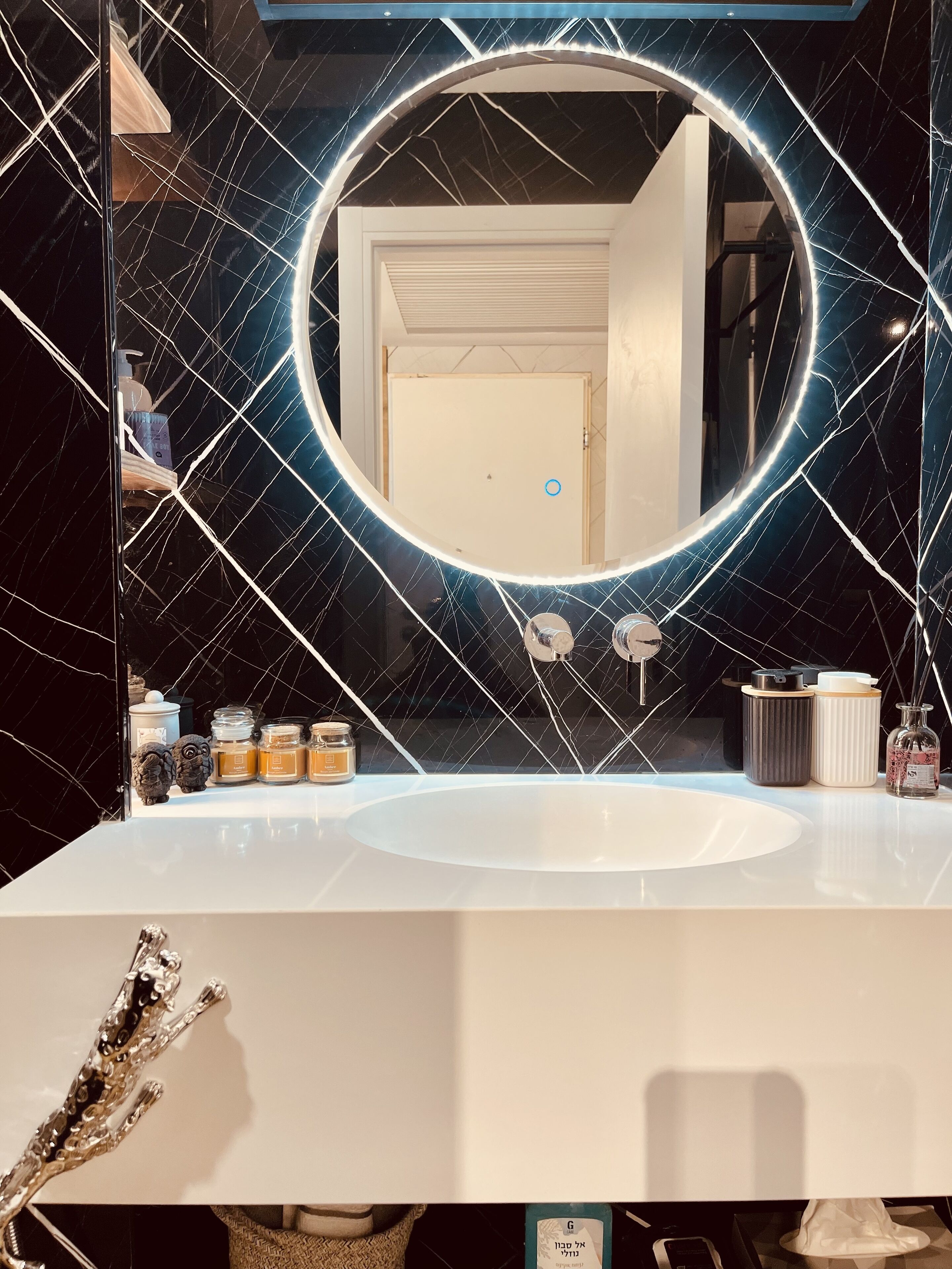 A bathroom with a white sink, a round mirror with a light, and a door in the background.