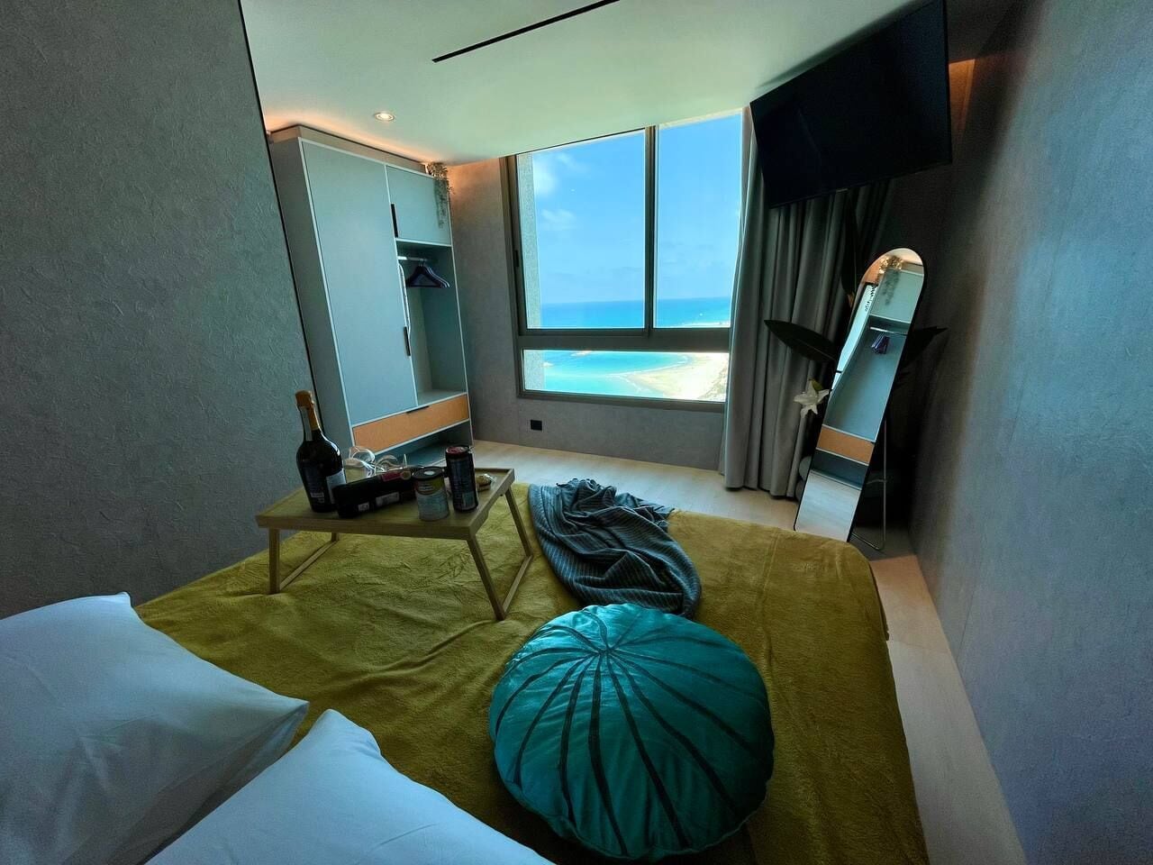 Una habitación de hotel con cama, una mesita con una botella y latas, un ventanal con vistas a la playa y un espejo.