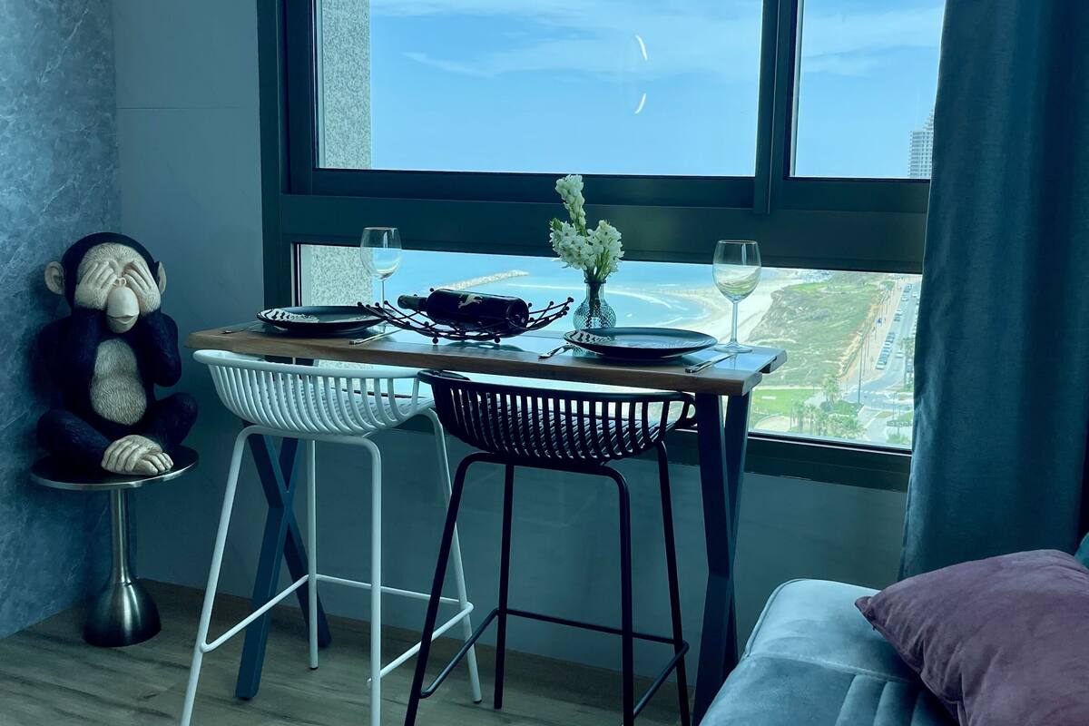 A cozy room with a large window, a small table with a vase of flowers, a teddy bear, and a book on a couch.