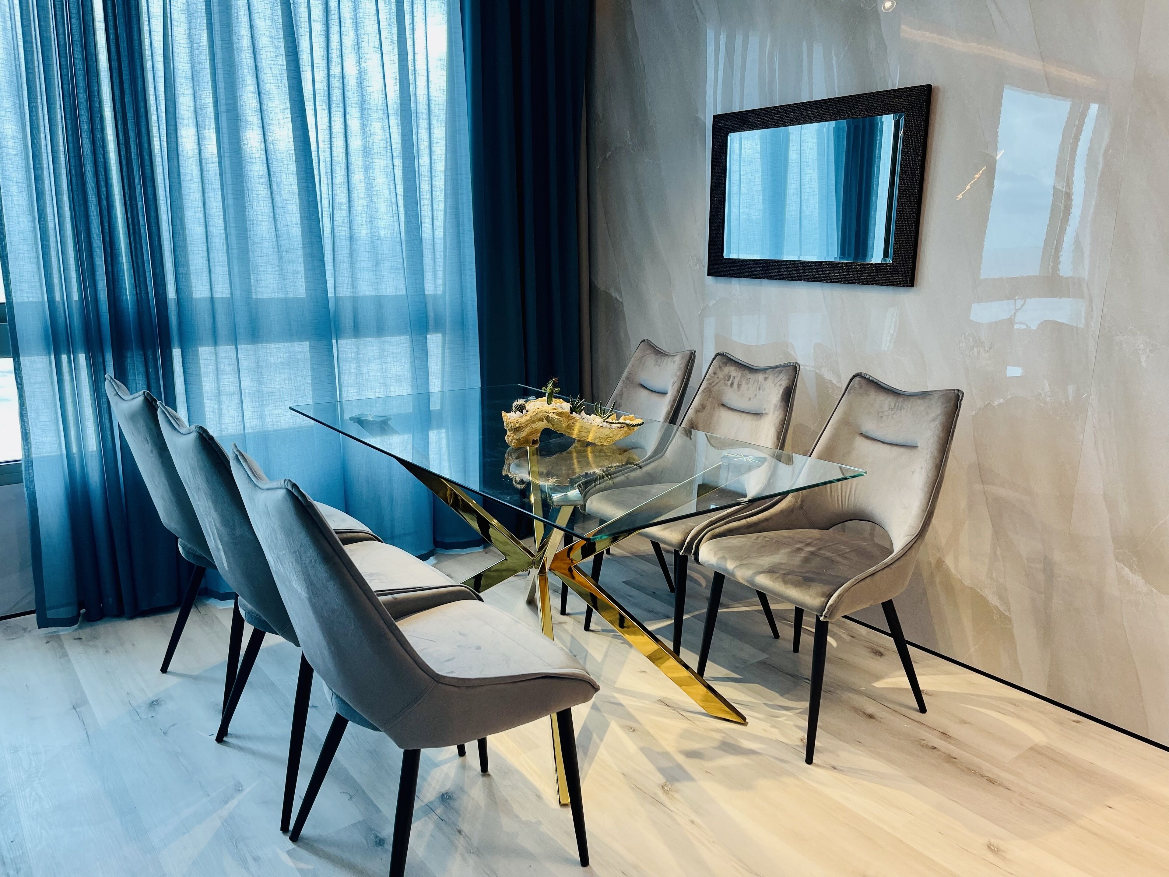 Een moderne eetruimte met een glazen tafel, omringd door zes stoelen, een vaas met bloemen en een grote spiegel aan de muur.