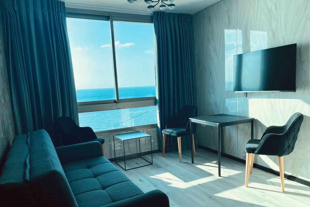 A modern living room with a sofa, a small table, and two chairs. A TV is mounted on the wall, and the room has large windows with curtains.