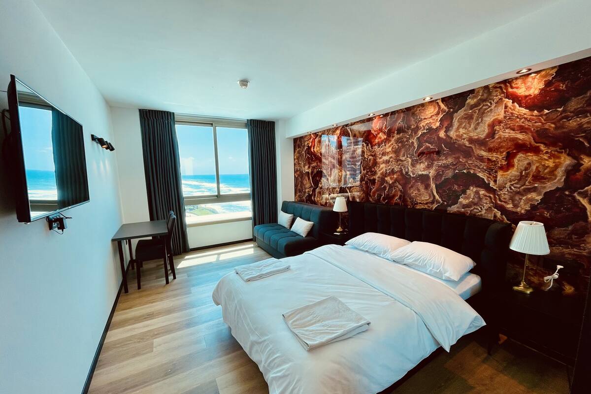 A modern hotel room with a large bed, a desk, a chair, and a TV. The room has a large window with a view of the ocean.