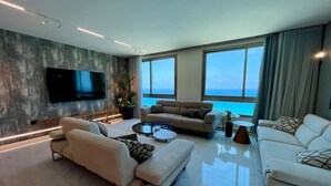A modern living room with a large flat-screen TV, two sofas, a coffee table, and a view of the ocean through large windows. - Carmel Holiday Apartments (Netanya)