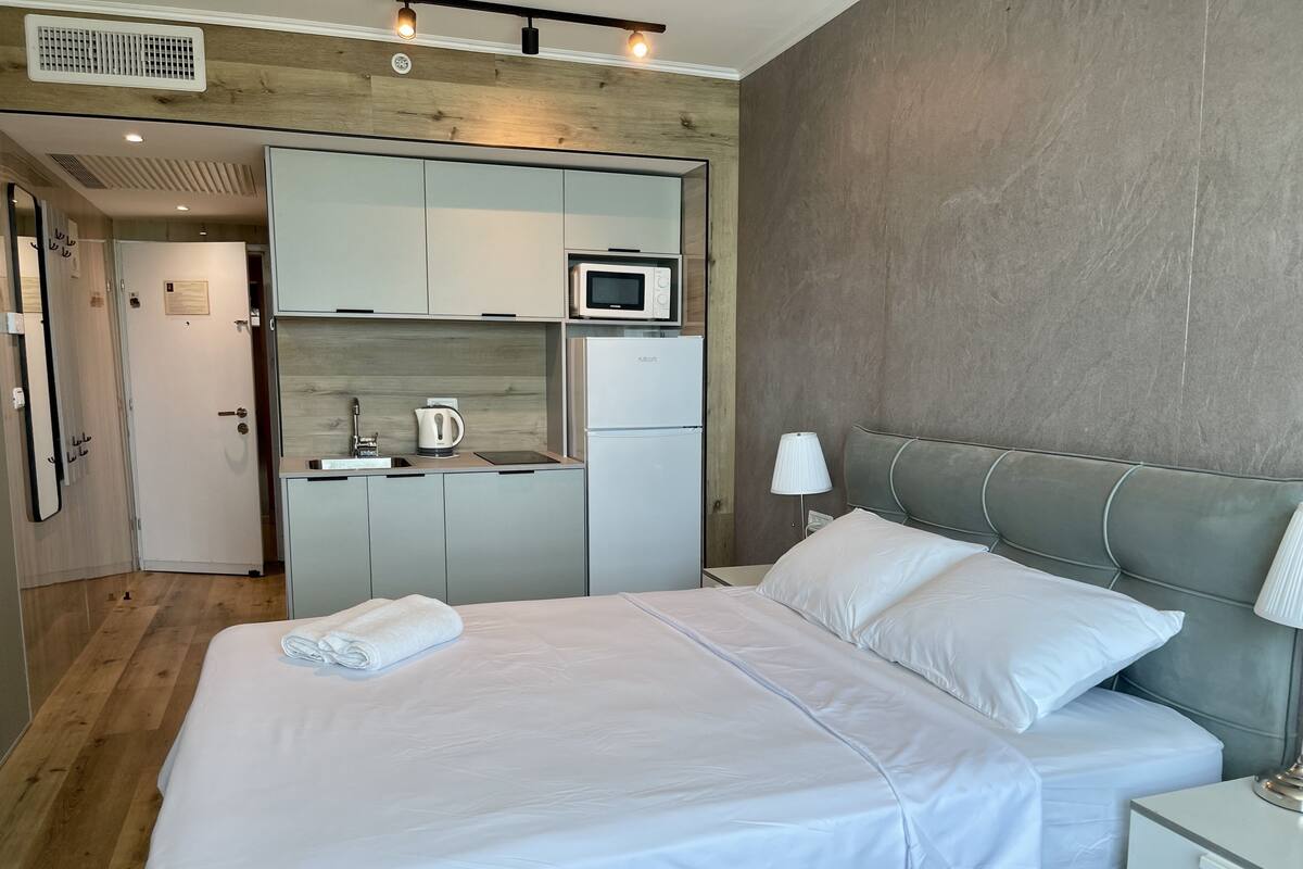 A modern hotel room with a bed, a nightstand, a lamp, a kitchenette with a microwave, a kettle, and a refrigerator.