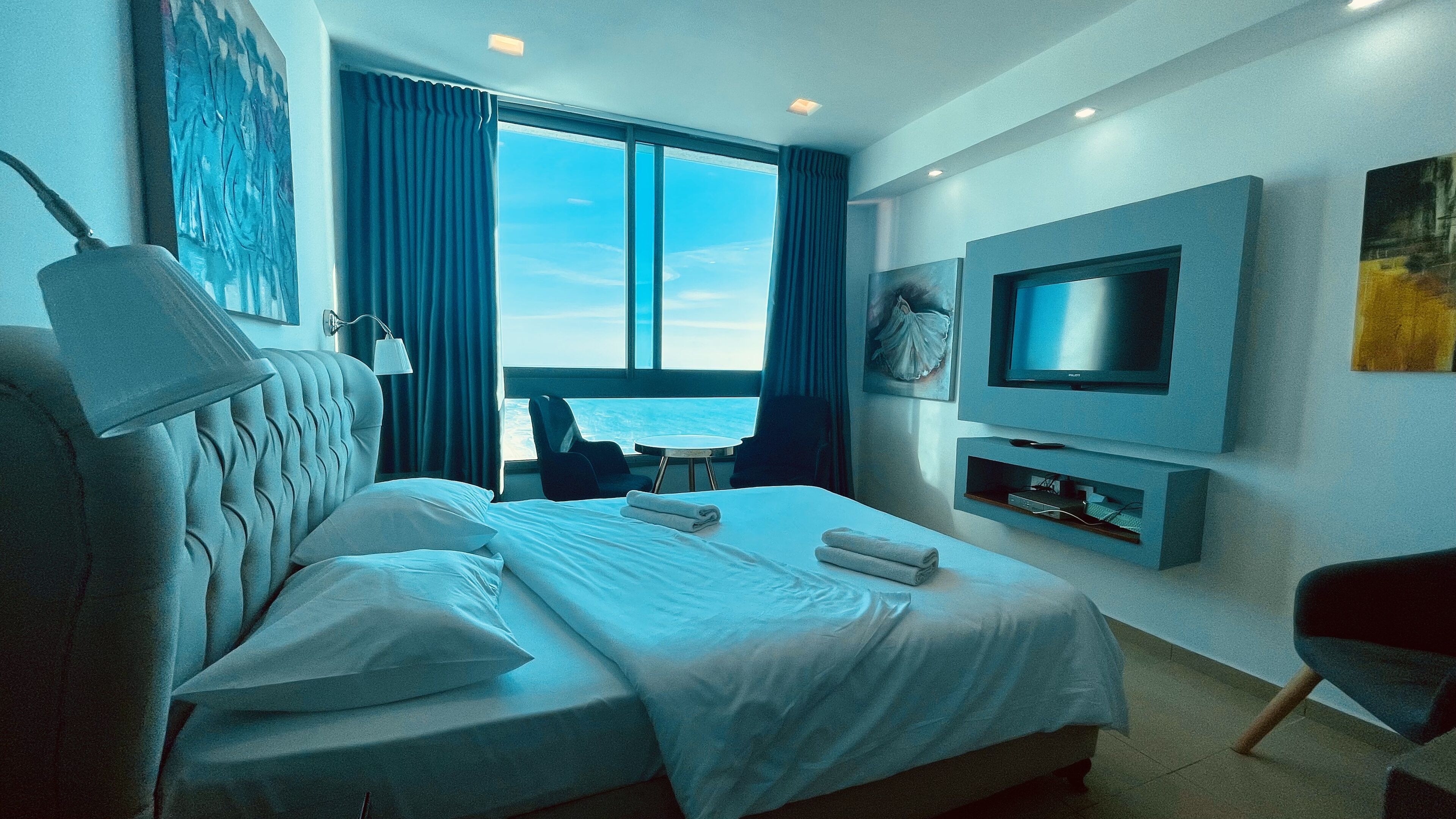 A modern hotel room with a large bed, a desk, a TV, and a view of the sea.