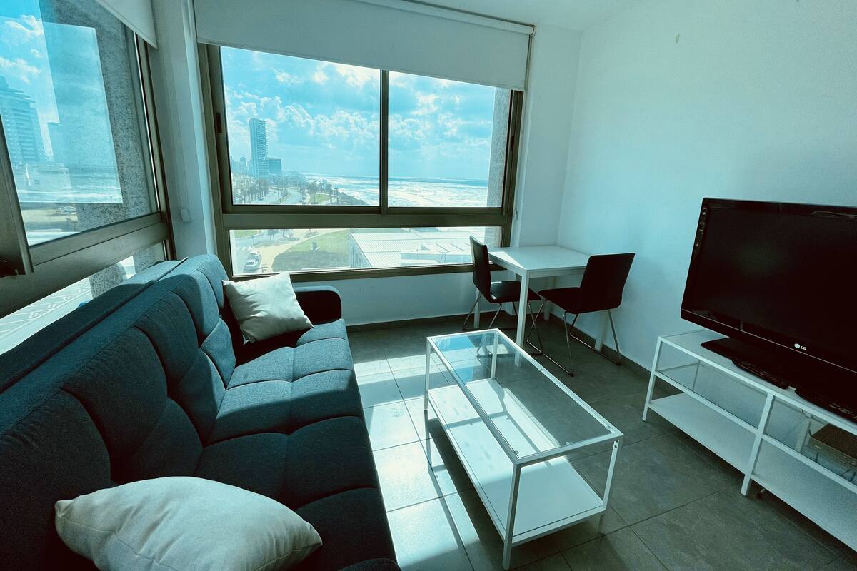 A modern living room with a sofa, a coffee table, a TV on a stand, and a large window offering a city view.