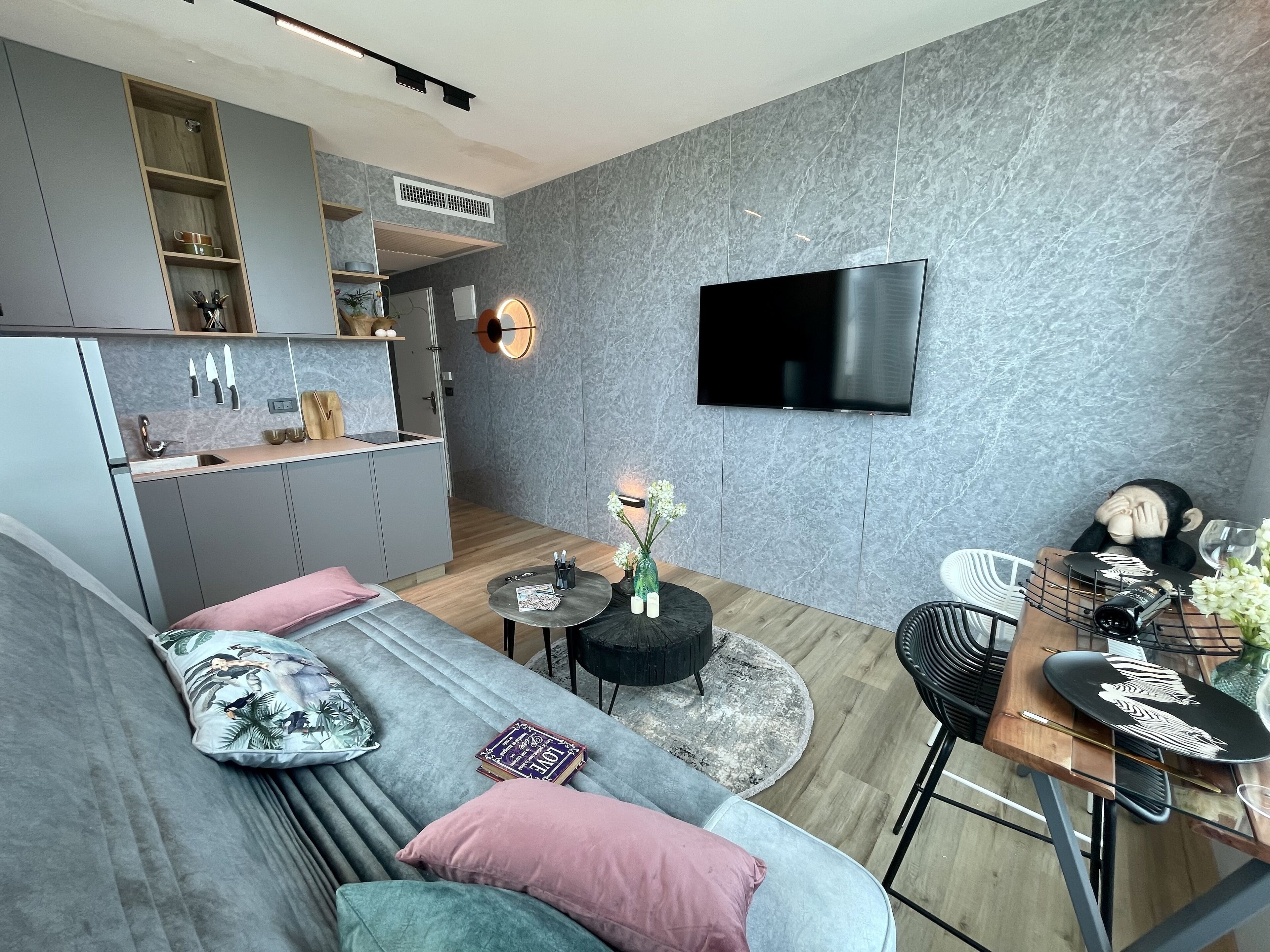 A modern living room with a grey sofa, a dining area with a wooden table, and a kitchen with grey cabinets.