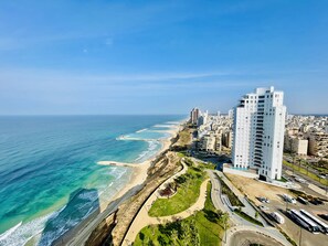 A coastal cityscape with a prominent white high-rise hotel, a beach, and a road curving along the coastline.