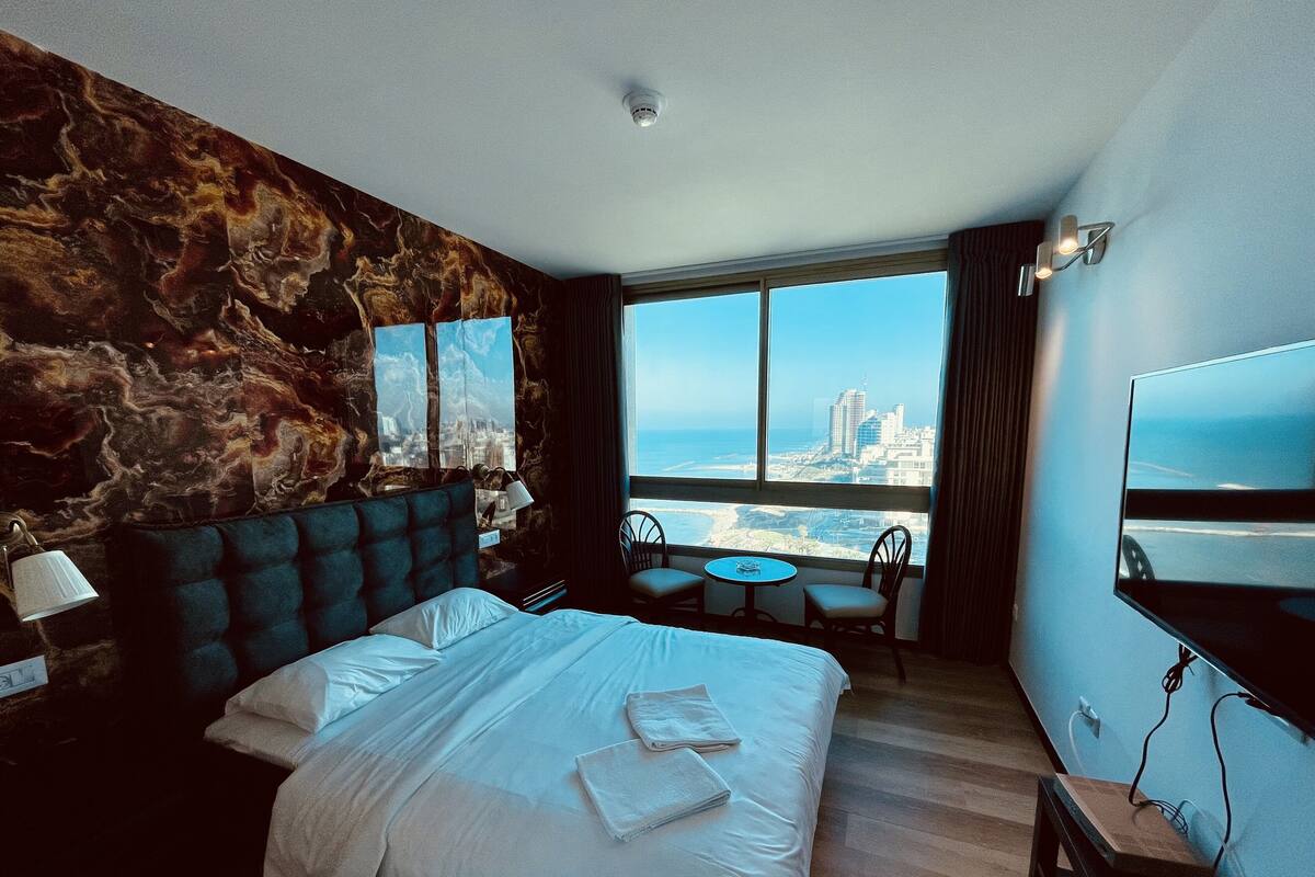 A hotel room with a large bed, a view of the city, and a small table with chairs.