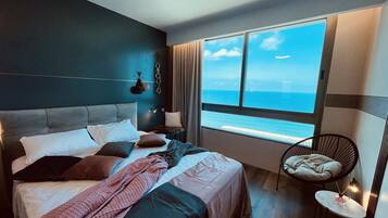 A modern hotel room with a large bed, a view of the sea, and a small table with a lamp.