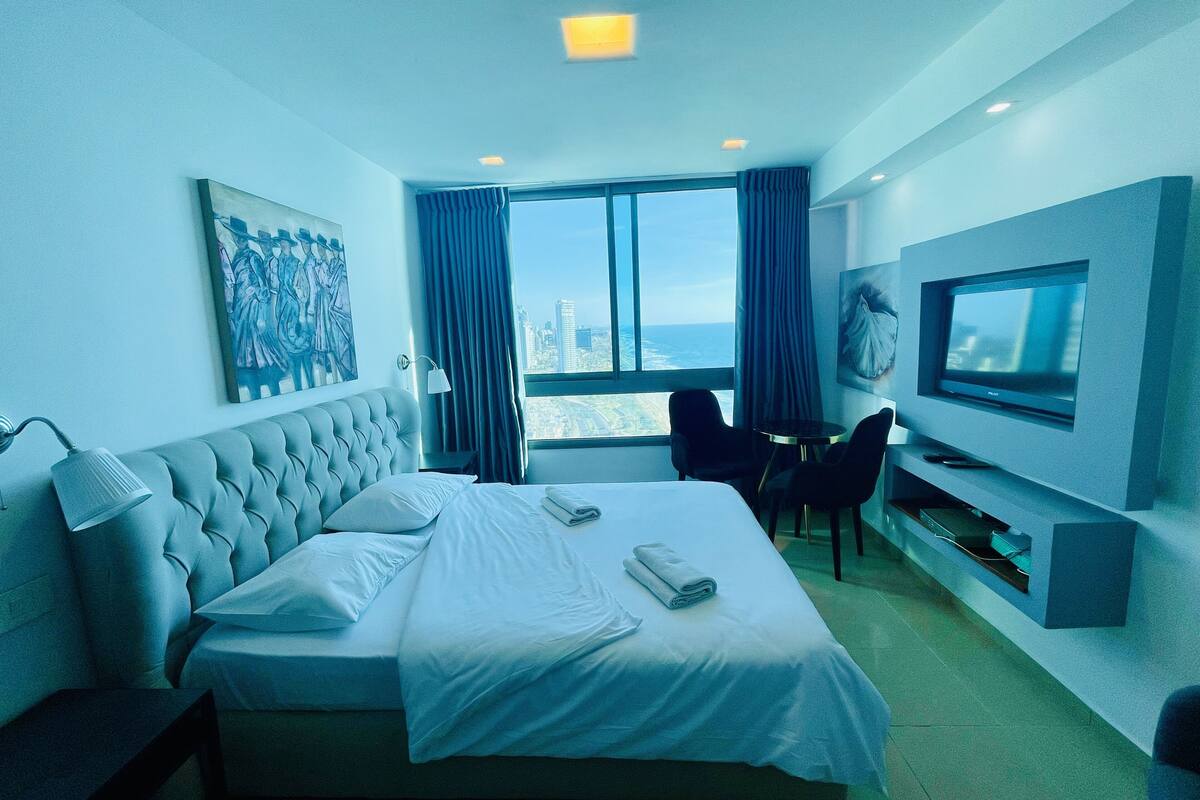 A hotel room with a large bed, a desk with two chairs, a flat-screen TV, and a city view through the window.