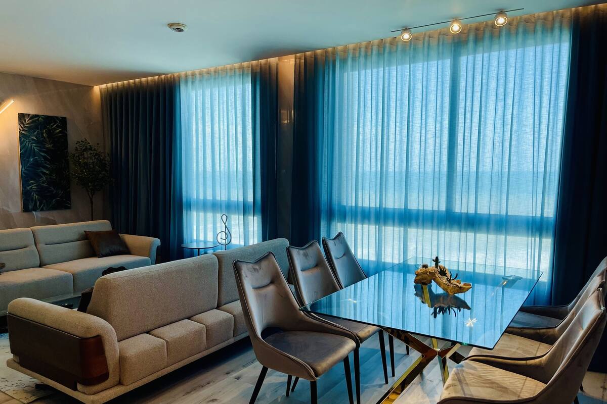 A modern living room with a glass dining table, leather chairs, a sofa, and large windows with blue curtains.