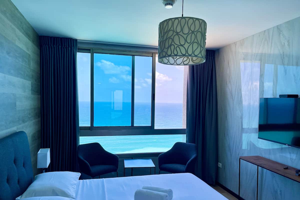 A hotel room with a bed, a desk, two chairs, a TV, and a large window with a view of the ocean.
