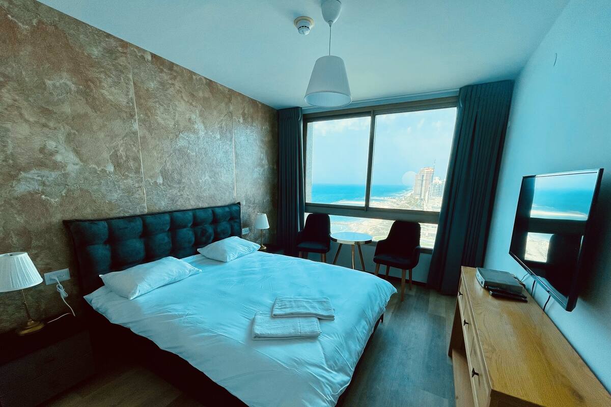 A hotel room with a bed, a desk, a TV, and a view of the beach.