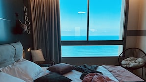 A hotel room with a large bed, a chair, a view of the ocean, and a window with curtains. - Carmel Holiday Apartments (Netanya)