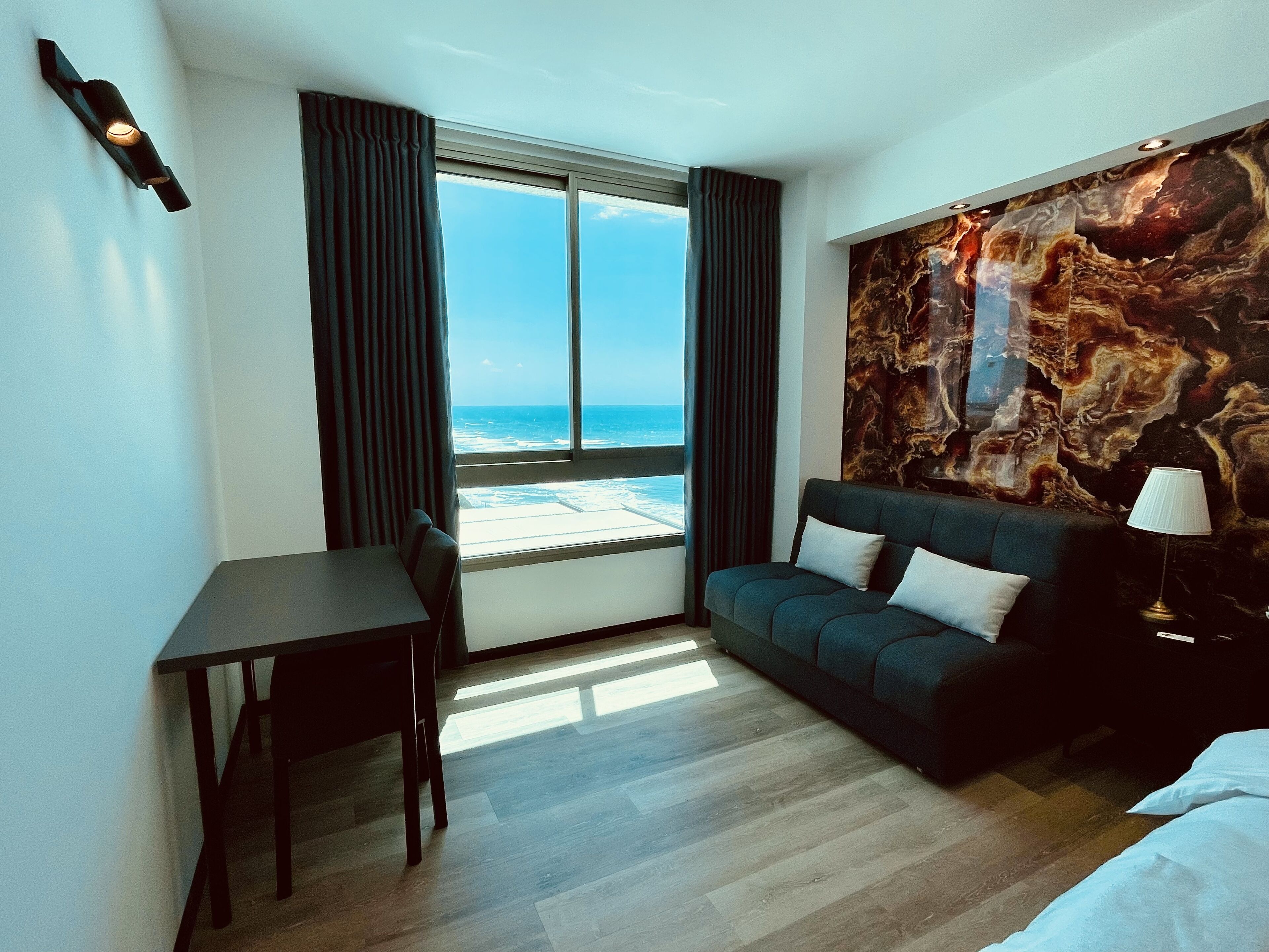Una habitación de hotel moderna con un ventanal grande que ofrece vistas a la playa, un sofá de color oscuro, una mesita y una lámpara.